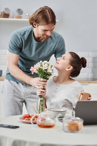 Gülümseyen genç bir adamın dikey portresi karısına sabah süprizi olarak çiçek veriyor. - Fotoğraf, Görsel