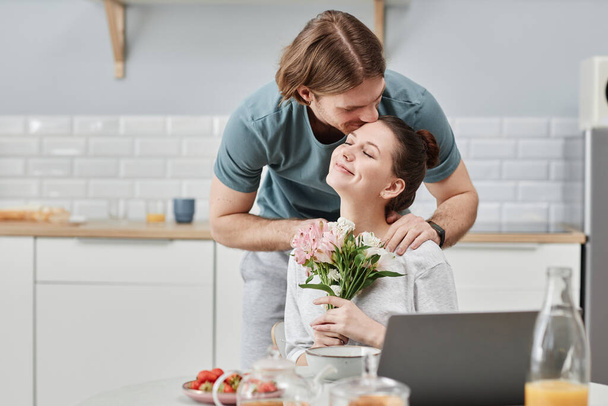 Portret van een glimlachende jongeman die 's morgens bloemen aan zijn vrouw geeft als verrassing en tedere kus op het voorhoofd, kopieer ruimte - Foto, afbeelding
