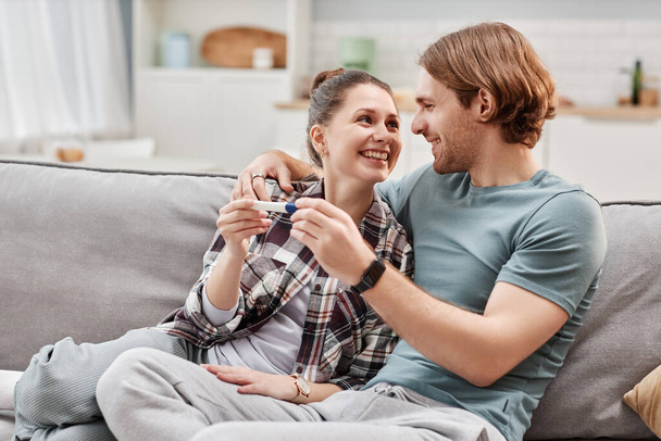 Portrét šťastného mladého páru držícího těhotenský test a objímající zatímco sedí na pohovce doma, kopírovat prostor - Fotografie, Obrázek