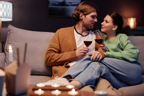 Retrato de pareja joven disfrutando de una velada romántica en casa y bebiendo vino abrazándose en el sofá, espacio para copiar - Foto, imagen
