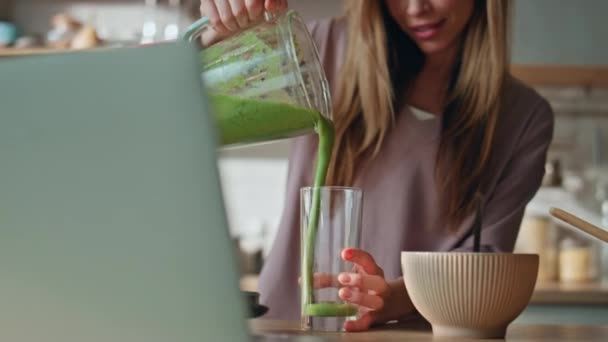 家庭用キッチンのノートパソコンカメラで料理ブログをレコーディングしている女性. 笑顔の魅力的な女の子は,グラスで滑らかな緑の野菜を注いで,コンピュータのウェブカムを見ています. ハッピー 主婦 料理 朝食. - 映像、動画