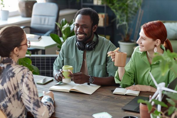 Многонациональная команда улыбающихся молодых бизнесменов, сотрудничающих в офисе, сидит за столом заседаний в зеленом офисе - Фото, изображение