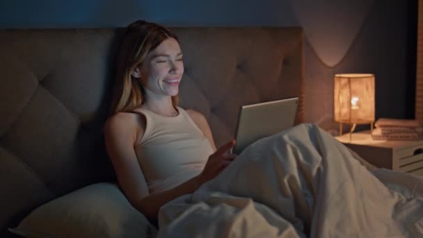 Κορίτσι βλέποντας ταινία στην οθόνη tablet στο άνετο κρεβάτι αργά το βράδυ. Χαμογελώντας χαλαρή γυναίκα απολαμβάνοντας αστείο βίντεο στον υπολογιστή pad κατά την κατάκλιση. Ευτυχισμένη όμορφη κυρία μηνυμάτων σε απευθείας σύνδεση πριν από τον ύπνο στο σκοτεινό υπνοδωμάτιο - Πλάνα, βίντεο