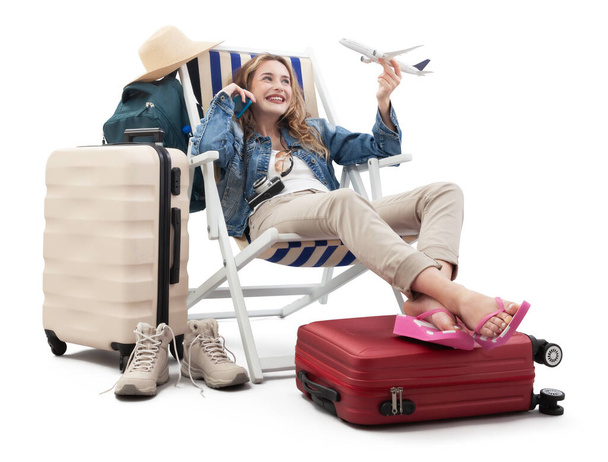 Tourist woman in travel attire, on deck chair with trolley suitcases, show a model airplane, using mobile phone. Summer beach holiday, flight and vacation travel booking. Travel influencer lifestyle - Photo, Image