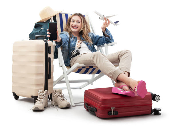 Tourist woman in travel attire, on deck chair with trolley suitcases, show mobile phone screen and model airplane. Summer beach holiday, flight and vacation travel booking. Travel influencer lifestyle - Photo, Image