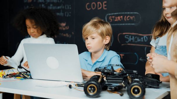 Κοντινό πλάνο του αγοριού που χρησιμοποιεί laptop προγραμματίζοντας κώδικα μηχανικής και το πρόγραμμα γραφής, ενώ η ομάδα των έξυπνων ποικιλόμορφων φοιτητών στέκεται περιβάλλεται από φίλο στην τάξη τεχνολογίας STEM στον πίνακα. Εκπομπή. - Φωτογραφία, εικόνα