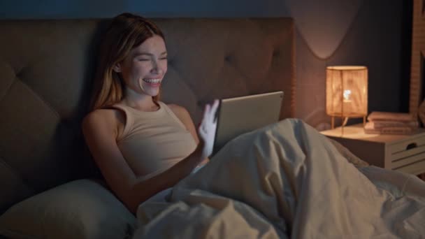 Sonriente chica tableta de mensajería acostado cómoda cama apartamento oscuro. Mujer atractiva feliz disfrutar de la comunicación en línea por ordenador de almohadilla en el dormitorio. Alegre dama viendo divertido vídeo en gadget digital. - Imágenes, Vídeo