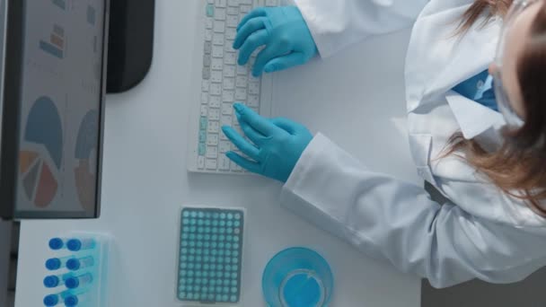 Ontwikkeling van het vaccin in het laboratorium. Technologisch modern laboratorium van medische analyses. Vrouw in witte jas zit achter de computer in beschermende handschoenen, typen op toetsenbord View from above. Hoog - Video