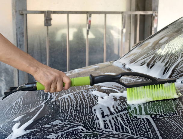 Bir kişi yeşil ve siyah bir fırçayla bir arabayı yıkıyor. Araba sabunlu suyla kaplıdır ve fırçayı fırçayla temizler.. - Fotoğraf, Görsel