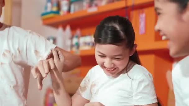 Handsome student playing with friend while put the clay on shirt at class. Group of diverse children working or modeling vase. Happy boy put water to young girl while laugh with happy. Edification - Footage, Video