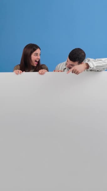 Vertikal. Junges erwachsenes Paar zeigt auf ein leeres Brett vor dem Hintergrund. Ratschläge kopieren Space-Attrappe. Mann und Frau zeigen eine Plakatwand vor blauem Hintergrund. Menschen präsentieren - Filmmaterial, Video