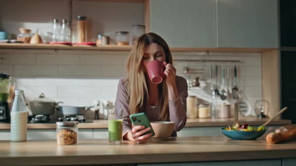 Unatkozó lány okostelefonnal kávézik a hangulatos konyhában. Pihentető, vonzó nő, aki a közösségi médiát nézi otthon. Komoly háziasszony élvezze forró ital a hétvégi konyha - Felvétel, videó