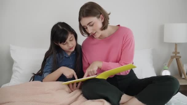 Caucasian mother reading story book to cute daughter listening story. Asian girl pointing while talking about story before bed time. Happy family sitting on bed while spend time together. Pedagogy. - Footage, Video