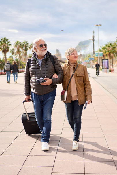Vertical. Couple of happy senior Caucasian tourist strolling hugging carefree with their luggage looking to the side. Pensioner people enjoying their journeys in a winter sunny day - Photo, Image