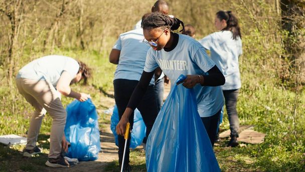 Afrika kökenli Amerikalı bir kız uzun pençe ve çöp torbalarıyla çöp topluyor, orman habitatını temizliyor ve bir grup gönüllüyle yasadışı çöplerle savaşıyor. Çöpleri toplayan aktivist. Kamera A. - Fotoğraf, Görsel