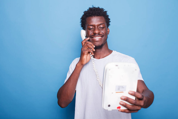 Young black man standing against isolated blue background is answering telephone call. Portrait of smiling african american guy enjoying a landline phone conversation with loved ones. - Photo, Image