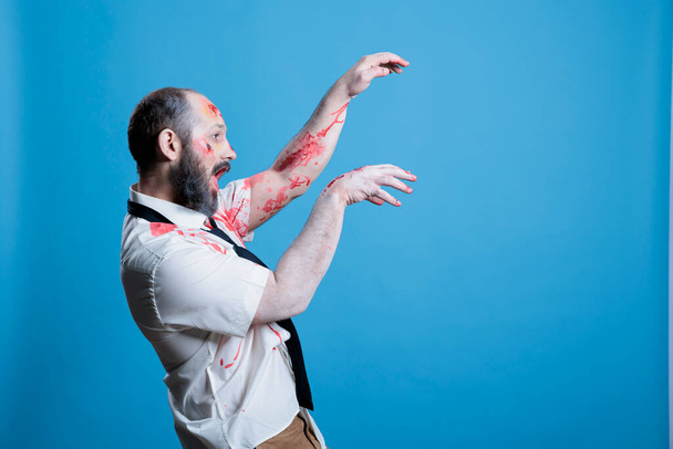 Possessed man covered in blood turned into zombie after being killed, haunting place. Wounded ghoulish cadaver limply walking towards victim, preparing for attack, studio background - Photo, Image