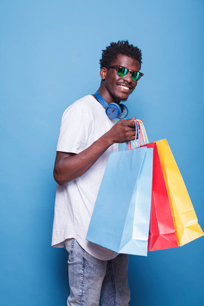 Αφροαμερικάνος που φοράει γυαλιά ηλίου, κρατάει τσάντες για ψώνια, και στέκεται μπροστά σε ένα απομονωμένο μπλε φόντο. Μαύρος άντρας με ασύρματα ακουστικά στο λαιμό, κουβαλάει πολύχρωμα δέματα. - Φωτογραφία, εικόνα