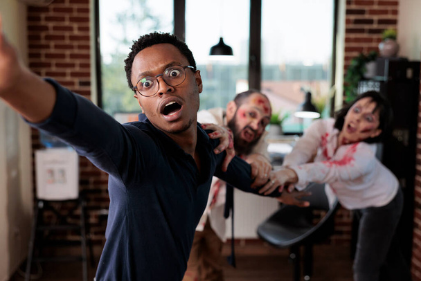 Közelkép afro-amerikai üzletemberről, akit ijesztő élőhalott szörnyek ragadtak el a cég irodájában. Fókuszálj az apokalipszis túlélő húzta agy eszik zombik elmosódott háttérrel - Fotó, kép