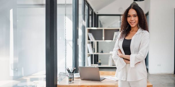 La confiada empresaria afroamericana se levanta con los brazos cruzados en un ambiente de oficina elegante, con su computadora portátil en el escritorio detrás de ella. - Foto, imagen