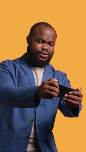 Vertikální video BIPOC osoba hrající videohry na smartphonu, s negativním výbuchem emocí po porážce. Frustrovaný hráč prohrává hru na telefonu, izolované přes studio pozadí, kamera B - Záběry, video