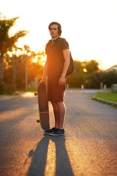Portrait, man and skateboard in outdoor, street and backpack on headphones, summer and sunset. University student, physical activity and sports in urban, skater and athletic in edgy, exercise and fun. - Photo, Image