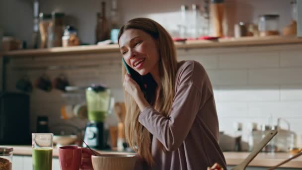 Μια οικιακή γυναίκα μιλάει στο κινητό της στην κουζίνα του διαμερίσματος από κοντά. Χαμογελώντας χαλαρή κορίτσι καλώντας smartphone πίνοντας πρωινό καφέ μόνο στην κουζίνα. Ευτυχισμένη ξέγνοιαστη νοικοκυρά μιλώντας κινητό τηλέφωνο στο σπίτι - Πλάνα, βίντεο