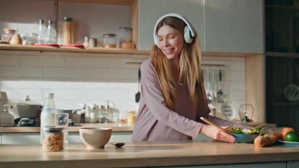 Kulaklıkçı kız, modern mutfağın içinde salata karıştırıp müzik dinliyor. Kulaklıklı kaygısız kadın sabah dairesinde sağlıklı bir kahvaltı hazırlıyor. Ev mutfağında granola pişiren mutlu bayan.. - Video, Çekim