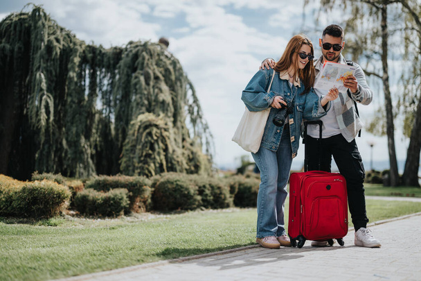Pareja de turistas en un viaje consultando un mapa juntos en un soleado parque de la ciudad, con equipaje a mano. - Foto, Imagen