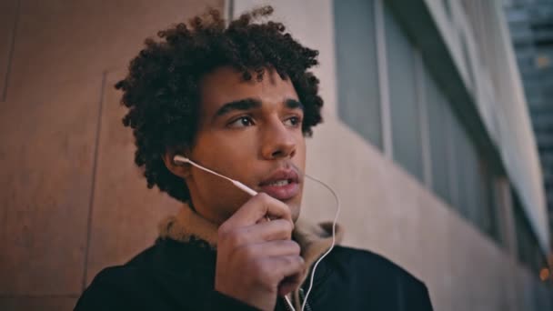 Handsome teenager talking headphones in urban setting closeup. Young latin american guy speaking in earphones at evening ambiance. Relaxed male hipster using headset for communication on dusk street. - Footage, Video