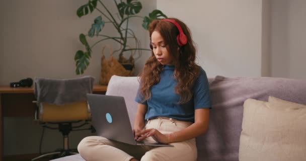 Latino nainen kuulokkeet toimii kannettavan istuu sohvalla. Latinalainen freelancer tietokoneella kääntyy pään sivuun miettien tehtävää. Sekalainen rotu toleranssi - Materiaali, video