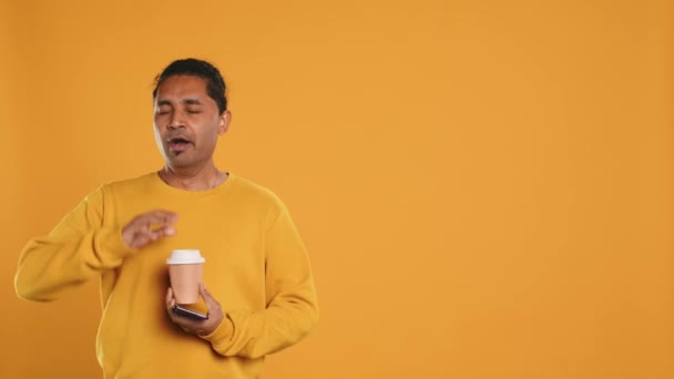 Indiai férfi élvezi kávé eldobható papír pohár és szöveges üzeneteket küldeni okostelefonon. Forró italok fogyasztása és csevegés barátokkal mobiltelefon, stúdió háttér, A kamera - Felvétel, videó