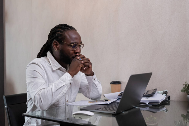 Der junge afroamerikanische Geschäftsmann fühlt sich gestresst und müde, während er von zu Hause aus mit Dokumenten arbeitet, die neben ihm gestapelt sind.. - Foto, Bild