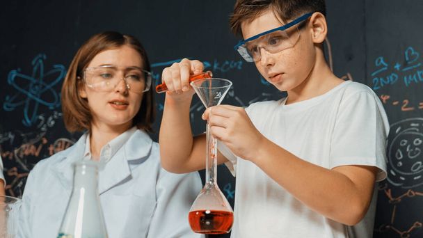 Close up of caucasian boy mixing chemical liquid while teacher giving advice. Instructor wearing lab suit looking for diverse student at table with beaker filled with colored solution. Erudition. - Photo, Image