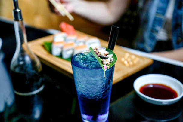 Стакан синей жидкости, расположенный рядом с тарелкой суши, демонстрирующий заманчивую комбинацию вкусов. - Фото, изображение