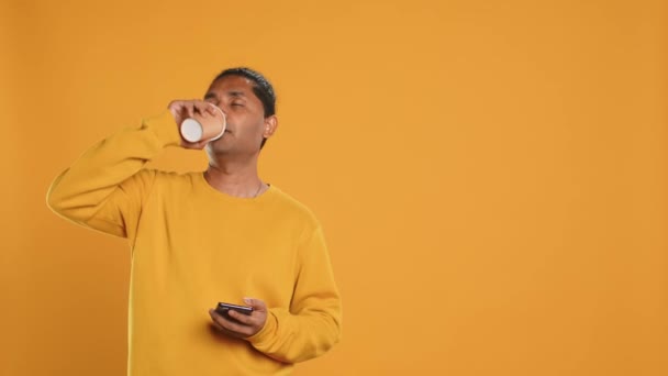 Indian man enjoying fresh coffee from disposable paper cup early in the morning to wake up and be energized. Person drinking hot beverage from recycled takeaway cup, studio background, camera A - Footage, Video