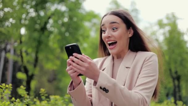 Young attractive woman cheering emotionally while holding mobile telephone and going by green pedestrian area. Charming woman bending hand with clenched fist while winning prize in online lottery. - Footage, Video