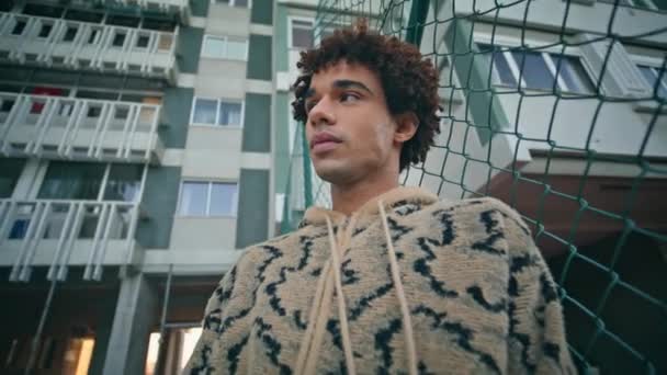 Casual student relaxing at street in textured sweater exuding calmness closeup. Relaxed latin american guy standing at urban fence at evening. Serious curly man posing at modern residential buildings - Footage, Video
