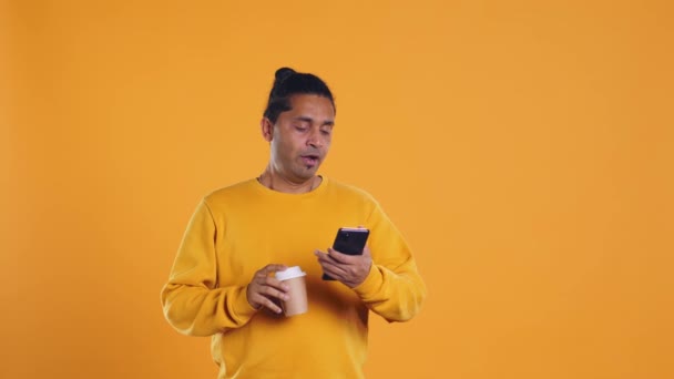Indian man enjoying coffee from disposable paper cup and send text messages on smartphone. Person drinking hot beverage and chatting with friends on mobile phone, studio background, camera B - Footage, Video