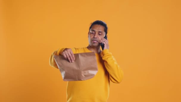 Man packing food in thermal backpack, fulfilling clients orders and answering phone ringing, studio backdrop. Indian person doing delivery to customers, discussing with them on cellphone, camera B - Footage, Video