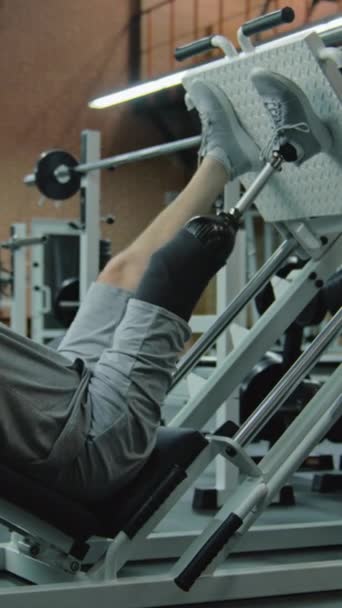 Κατακόρυφη βολή αθλητή με προσθετικά τραίνα ποδιών στο μηχάνημα τύπου ποδιών στο σύγχρονο γυμναστήριο. Ενηλίκων αθλητικός άνθρωπος με τεχνητό άκρο εκτελεί ασκήσεις δύναμης χρησιμοποιώντας επαγγελματικό αθλητικό γυμναστήριο - Πλάνα, βίντεο