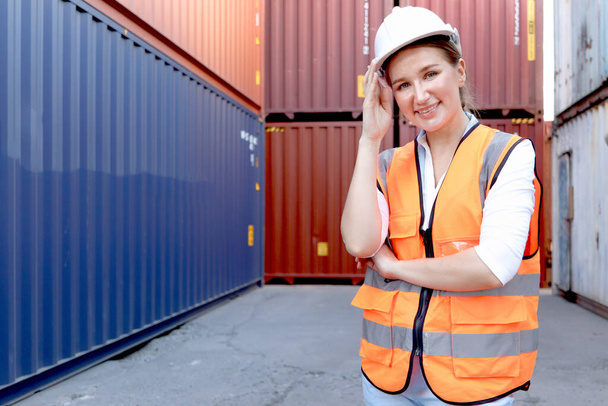 Portret van vrolijke glimlachende jonge mooie vrouw ingenieur baas met blond haar dragen veiligheidsvest en witte helm, staan met vertrouwen arm gekruist op logistieke scheepvaart containerwerf. - Foto, afbeelding