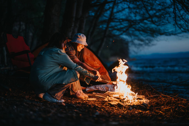 Kaksi ystävää paistaa vaahtokarkkeja yli viihtyisä kokko järven hämärässä, esittelee rauhallinen camping kokemus. - Valokuva, kuva