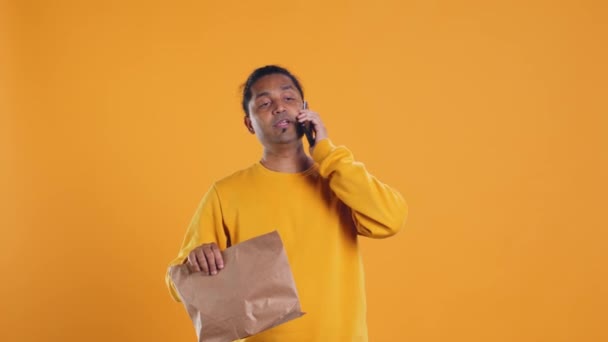 Indian man holding thermal backpack answering phone, fulfilling clients orders, isolated over studio background. Food delivery service employee talking with customer during telephone call, camera B - Footage, Video