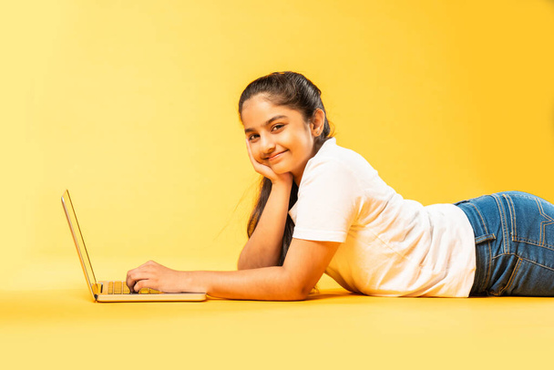 Indian asian small girl using laptop while lying on yellow floor and background isolated - Photo, Image