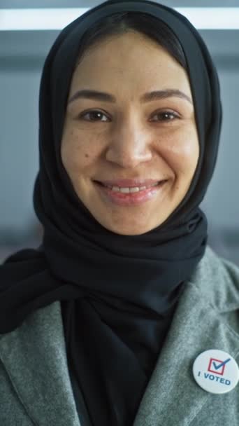 Tiro vertical de mujer árabe, votante de las elecciones de los Estados Unidos de América. La mujer se encuentra en un moderno centro de votación, posa, sonríe y mira a la cámara. Antecedentes con mesas de votación. Concepto de cívico - Metraje, vídeo