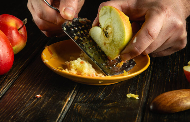 Appeldessert maken. De kok raspt een verse appel met een rasp in zijn handen op een keukenbord. - Foto, afbeelding