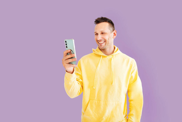 Joven sonriente alegre hombre caucásico 30s en sudadera con capucha amarilla haciendo foto selfie en el teléfono celular móvil post foto en la red social aislado en rosa fondo retrato de estudio. - Foto, Imagen