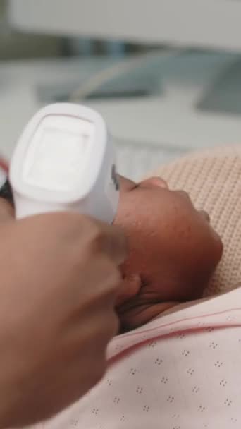 Vertical medium closeup of unrecognizable pediatrician checking temperature of little African American baby girl sleeping in mothers arms with infrared thermometer while having checkup appointment - Footage, Video