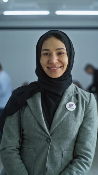Tiro vertical de mujer musulmana, votante de las elecciones de los Estados Unidos de América. La mujer se encuentra en un moderno centro de votación, posa, sonríe y mira a la cámara. Antecedentes con mesas de votación. Concepto de cívico - Metraje, vídeo